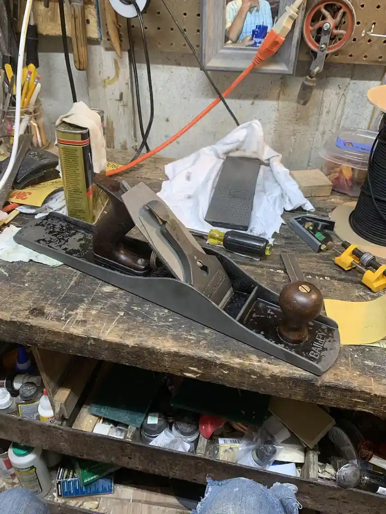 A well-worn woodworking bench is cluttered with tools, including a prominent Stanley Bailey hand plane, a sharpening stone…