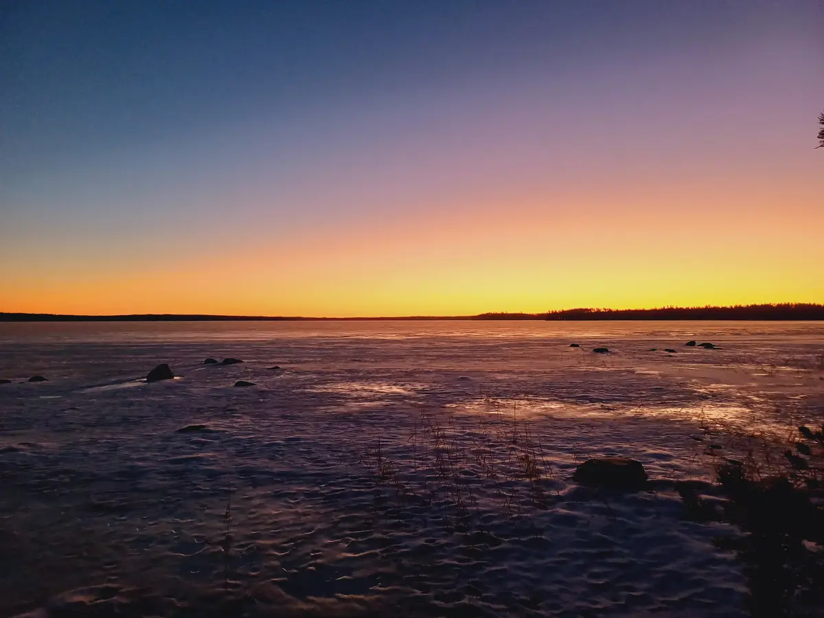 A serene winter sunset over a frozen lake, with vibrant orange and purple skies reflecting on the ice. Dark rocks and sparse…