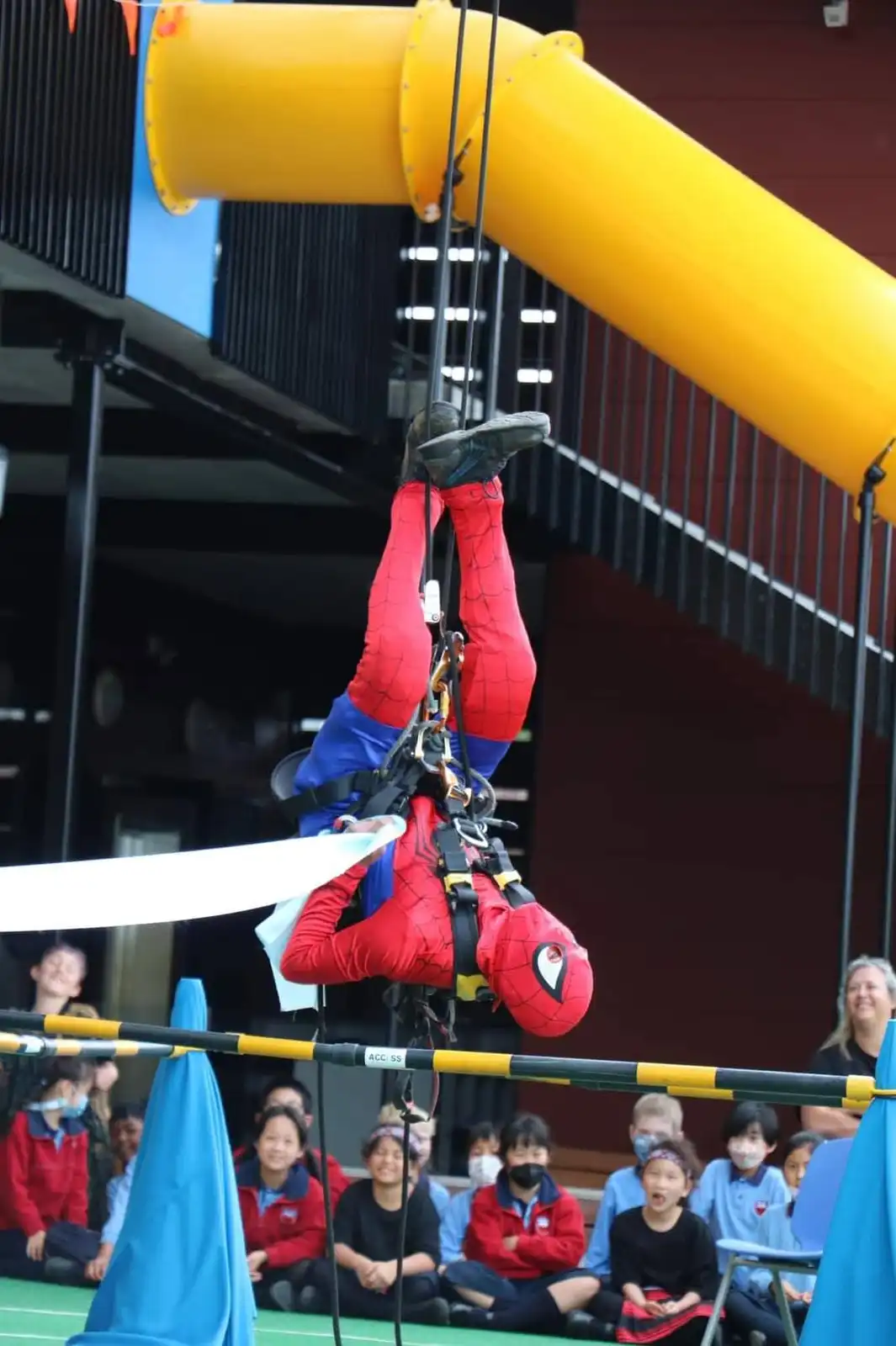 A person in a full Spider-Man costume is suspended upside down from a rope, hanging above a group of children seated on the…