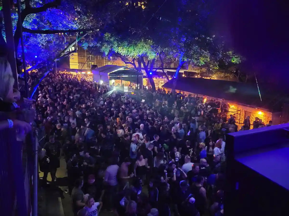 A high-angle nighttime photo of a densely packed outdoor concert crowd under colorful stage lights, with trees and buildings…