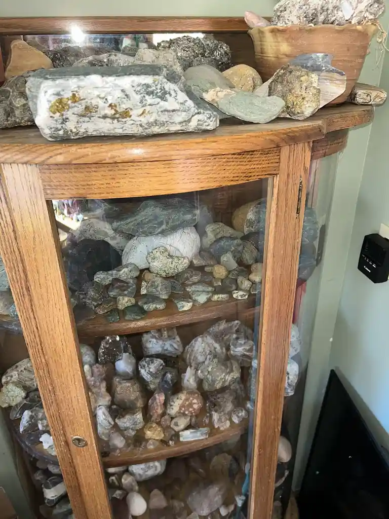 A wooden corner display cabinet filled with numerous rocks and minerals across multiple shelves, with more rocks piled on…