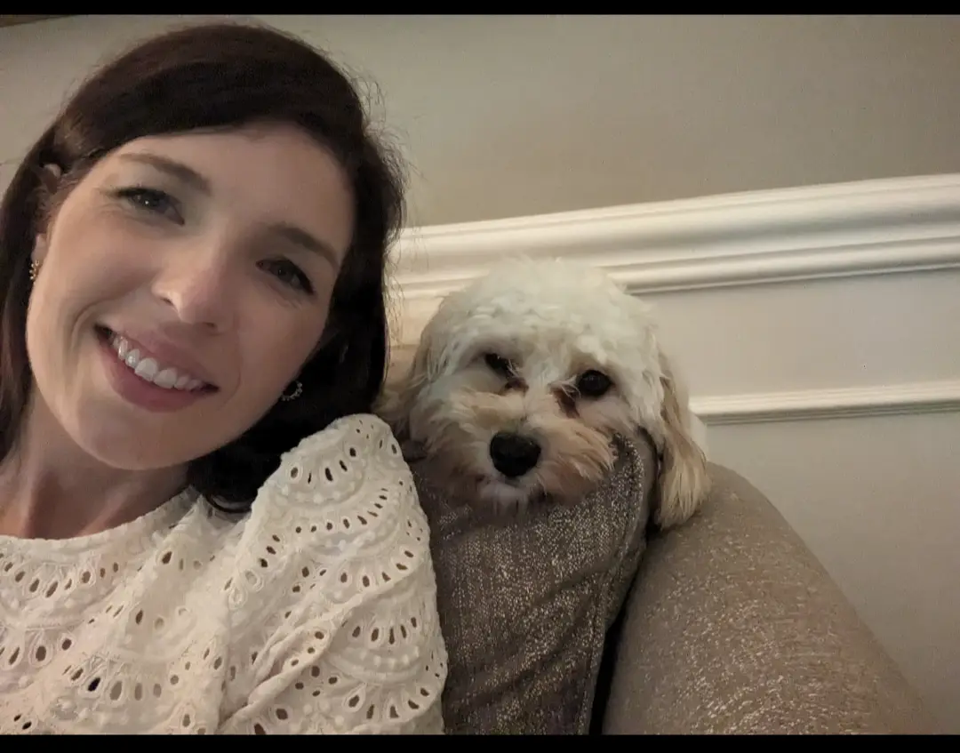 A woman with dark hair and a white lace top smiles at the camera while sitting on a couch next to a small, fluffy white dog…