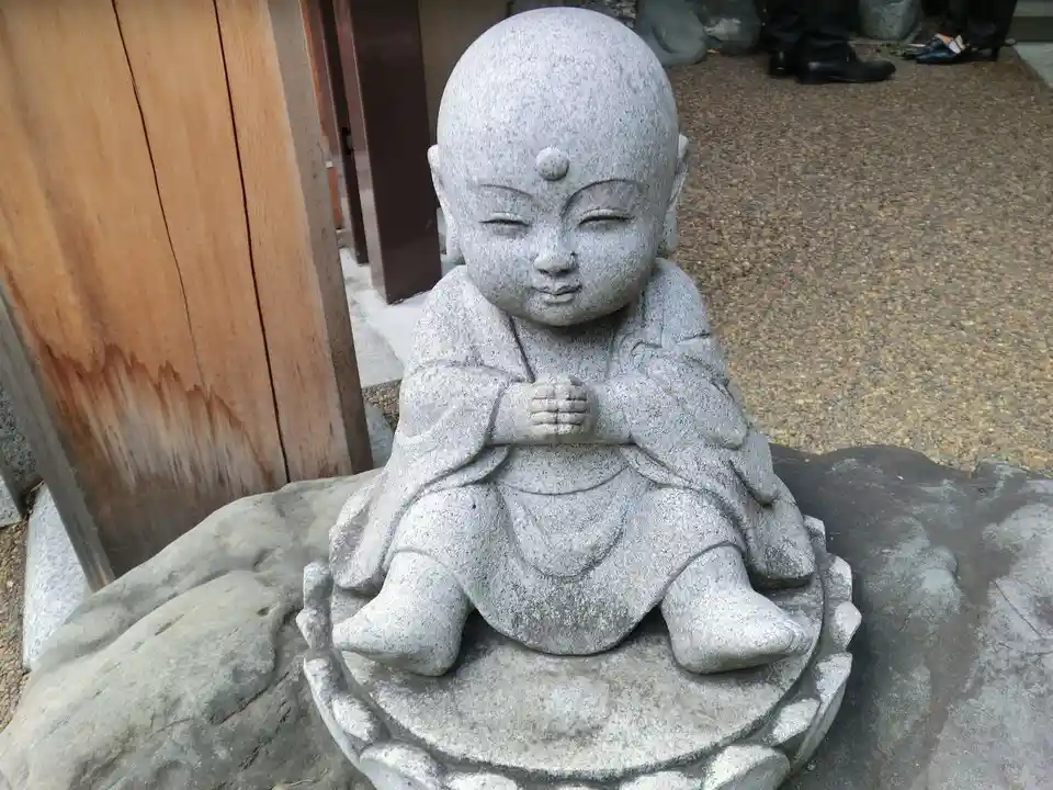 A stone statue of a smiling, bald, seated child monk with hands clasped, sitting on a lotus base outdoors. The overlaid text…