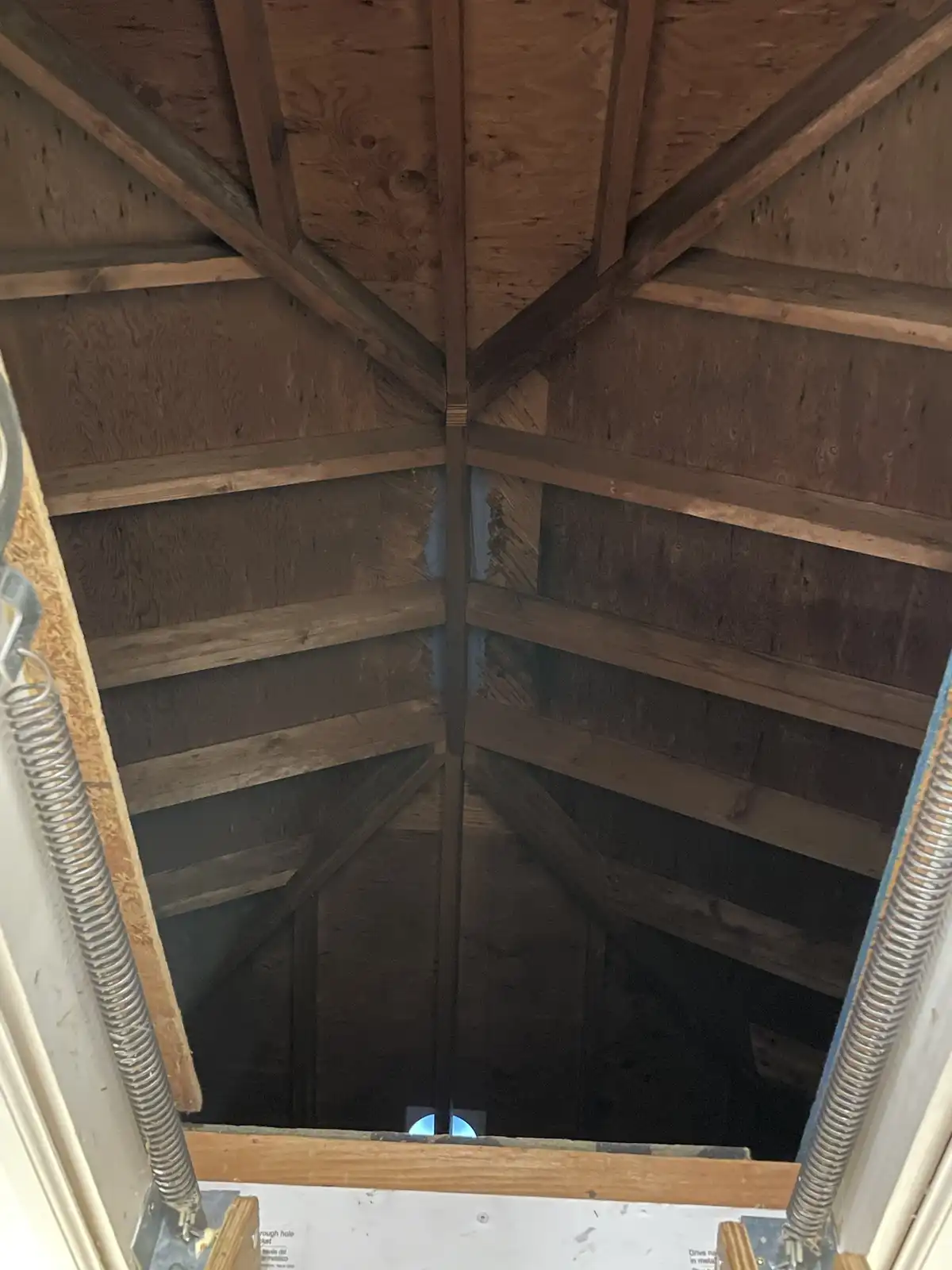 A photo looking up from inside an attic hatch into a dark, wooden roof structure with exposed beams and insulation. The…