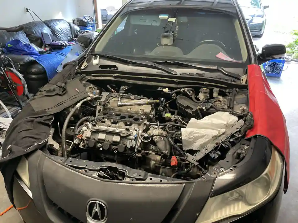 A black Acura car with its hood open, revealing a disassembled engine bay with tools and parts. A red cloth covers the…