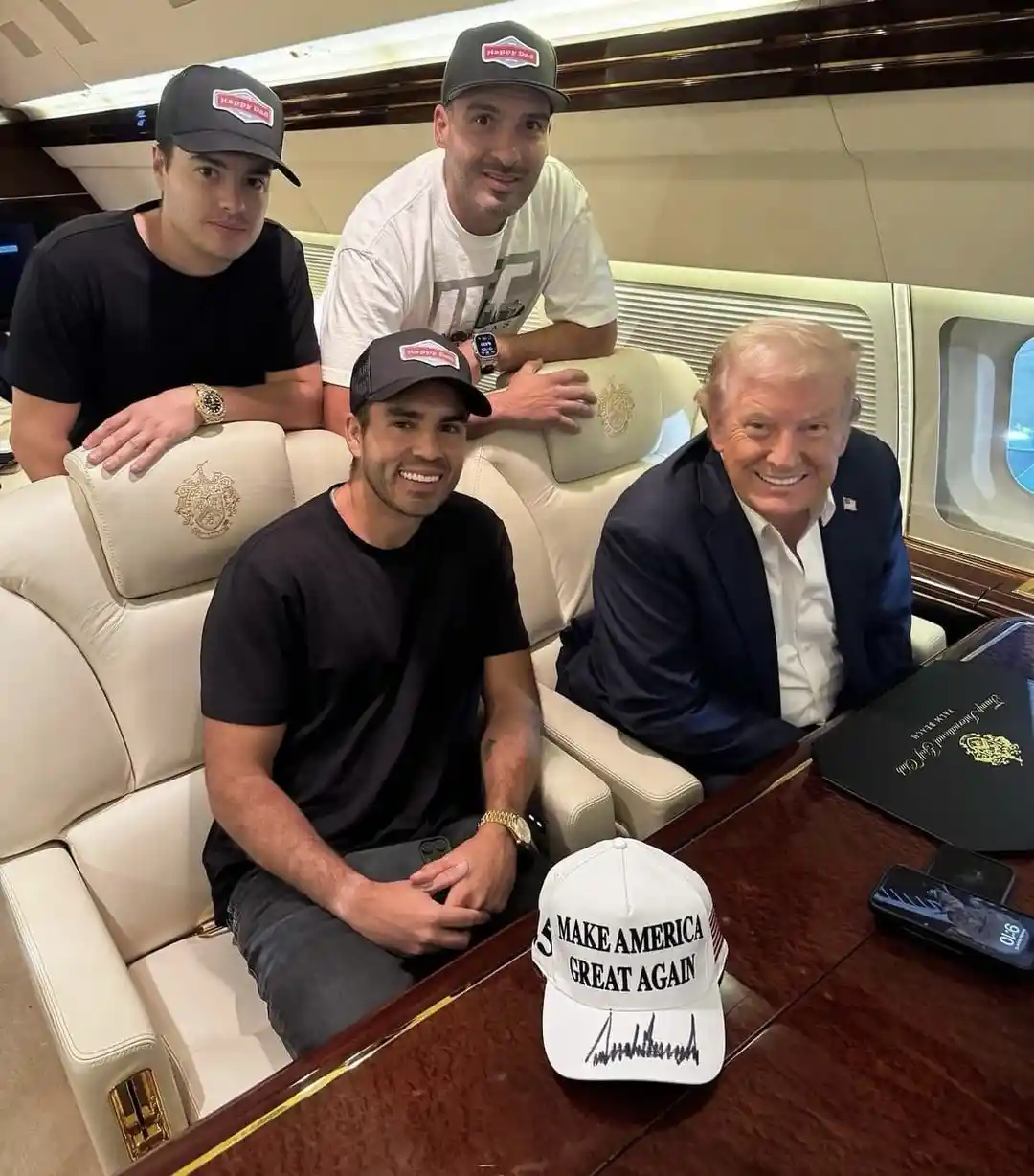 Four men are seated inside a luxurious private jet cabin, three wearing matching 'Happy Dad' hats and one wearing a suit. A…