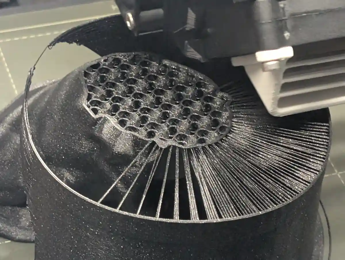 A close-up photo shows a 3d printer actively extruding black filament to build a hat-shaped object with a honeycomb pattern…