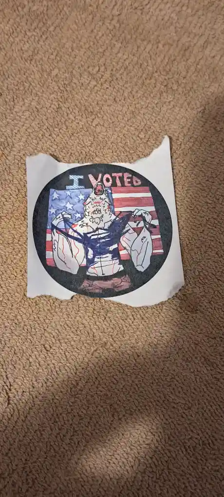 A hand-drawn, torn paper 'I VOTED' sticker lies on a brown carpet. The circular sticker features a muscular, cartoonish…