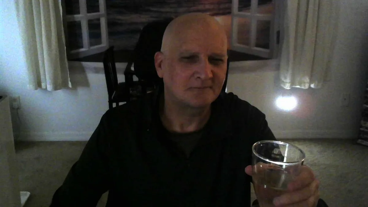 A bald man in a dark hoodie sits indoors holding a clear glass, looking slightly squinty-eyed and unimpressed. Behind him…