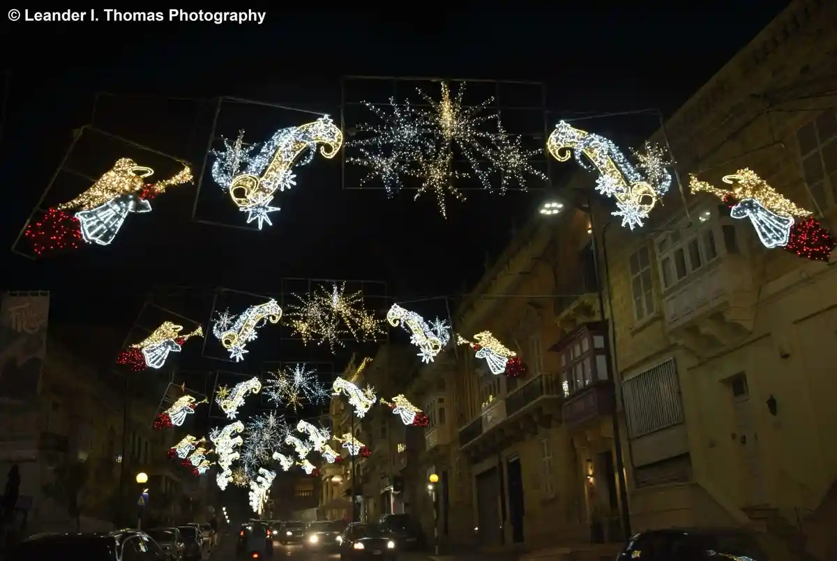 A nighttime street scene with elaborate, glowing Christmas decorations hanging overhead, including angels and starburst…