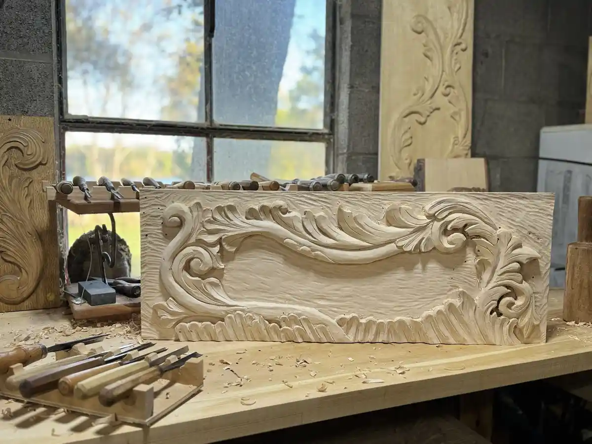 A close-up photo of an ornate, unfinished wooden carving on a workbench in a workshop, surrounded by carving tools and wood…