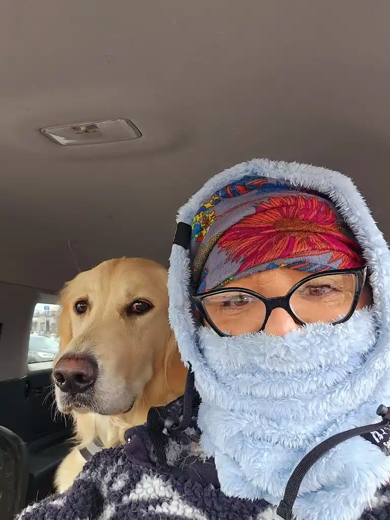 A person inside a car wearing a light blue fuzzy hooded scarf and glasses, with a golden retriever sitting beside them. The…