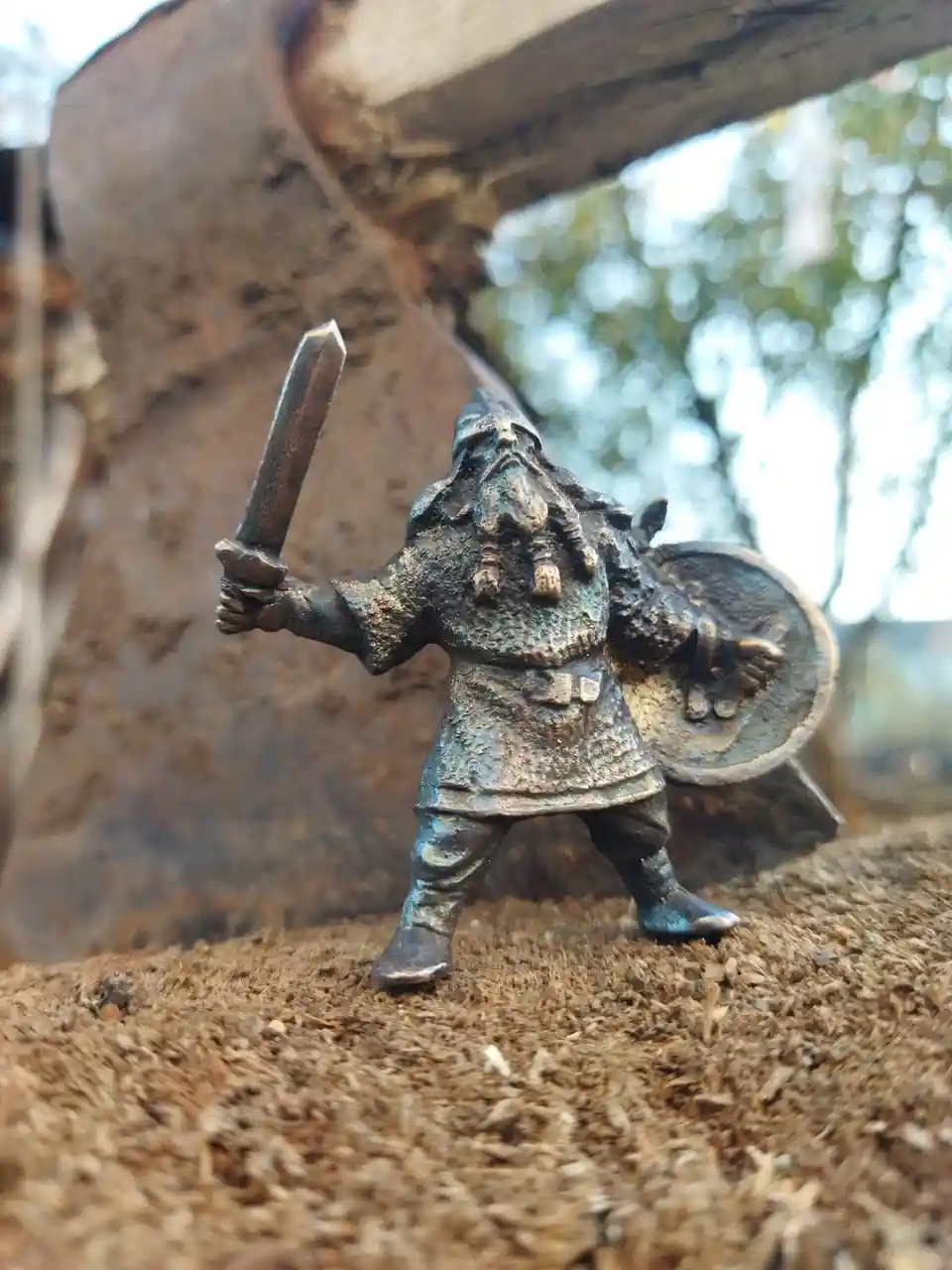 A small, bronze-colored figurine of a bearded warrior, resembling a dwarf or Viking, stands on wood shavings holding a sword…