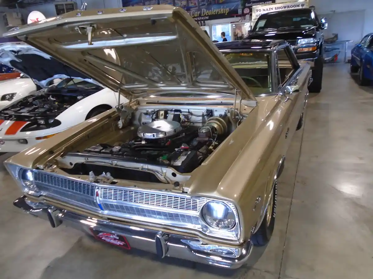 A tan classic American car with its hood open, revealing a chrome engine, is parked in a car dealership showroom with other…