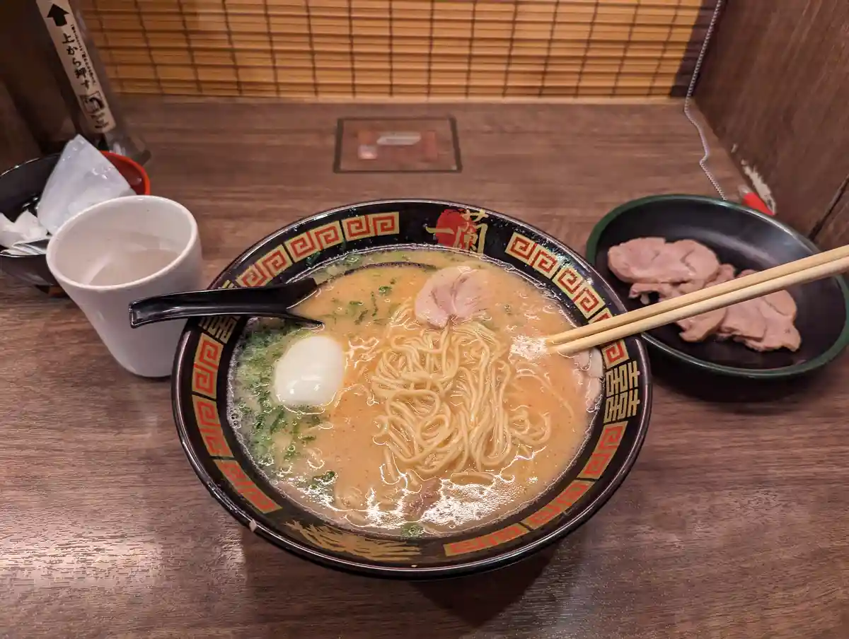 A close-up photo of a steaming bowl of ramen on a wooden table, featuring noodles, broth, a soft-boiled egg, and slices of…
