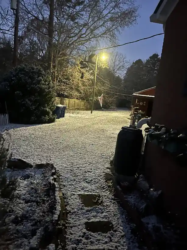 A snowy backyard at dusk, illuminated by a streetlamp, with a path leading toward a fence and shed. The ground is covered in…