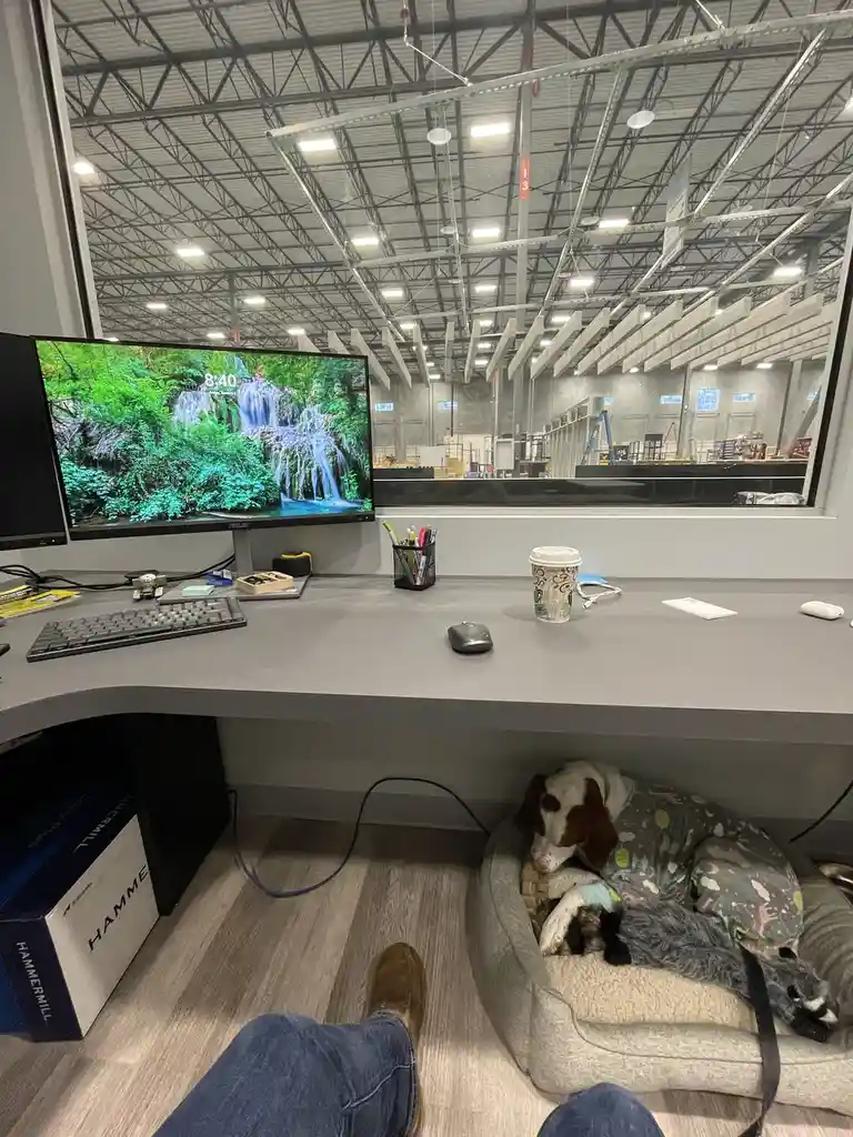 A person's perspective from a home office desk, showing a computer monitor displaying a waterfall wallpaper with the time…
