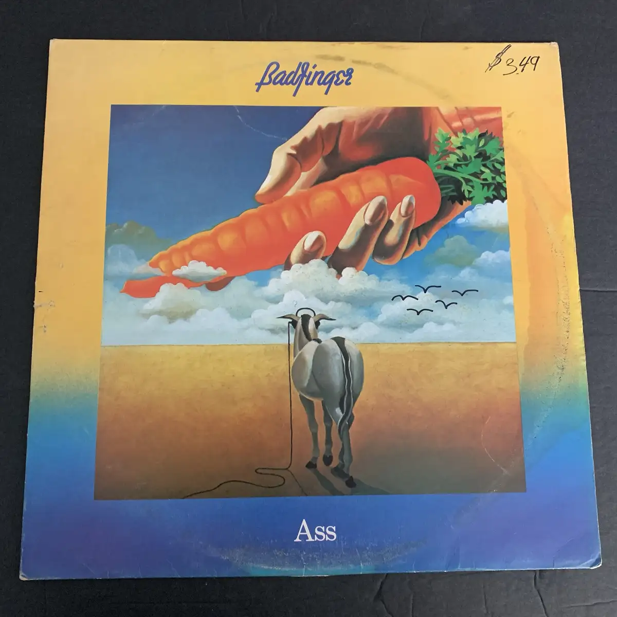 The image shows the album cover for Badfinger's 'Ass,' featuring a large hand holding a giant carrot above a field where a…
