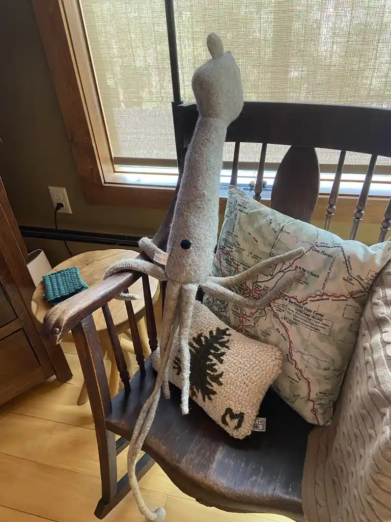 A handmade, light gray knitted octopus plush toy is draped over the back of a dark wooden rocking chair, its tentacles…