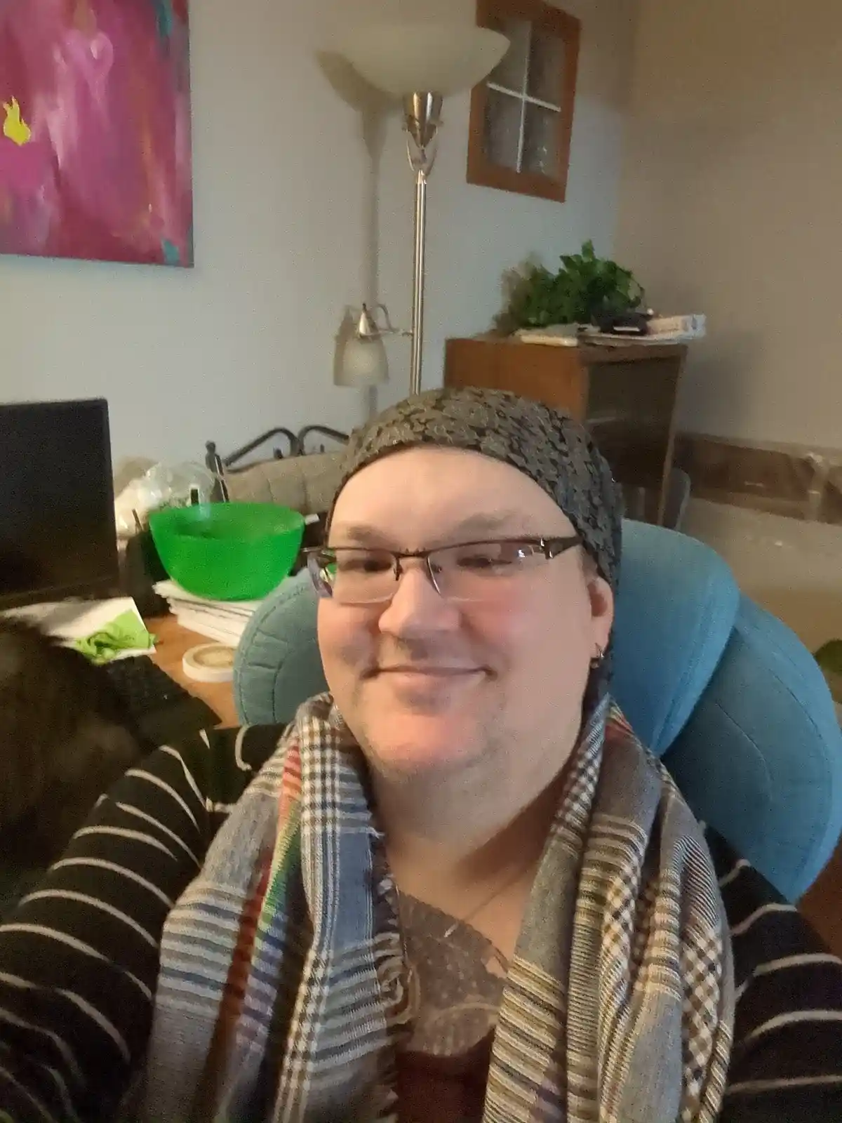 A person wearing glasses and a head covering sits in a blue chair in a home office, smiling subtly while surrounded by a…