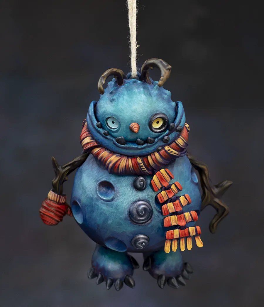 A whimsical, handcrafted blue monster ornament with large eyes, small horns, and a colorful striped scarf hangs by a string…