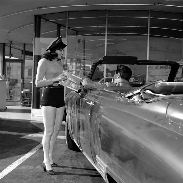 A vintage black-and-white photo shows a woman in a short skirt and hat handing a magazine to a man in a convertible car. The…