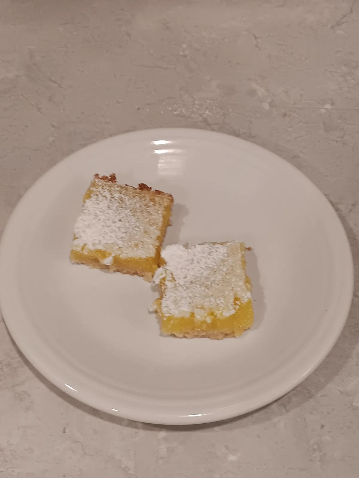 Two square lemon bars dusted with powdered sugar sit on a plain white plate, which rests on a light-colored, textured…
