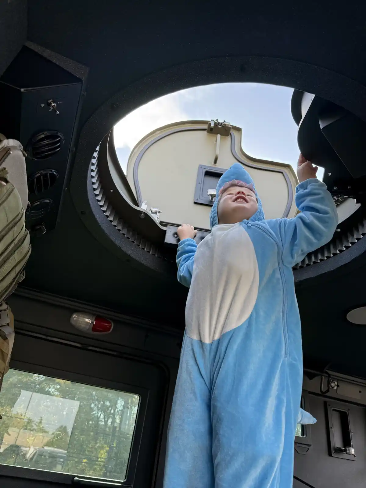 A young child wearing a blue shark onesie stands inside a military vehicle, looking up and smiling joyfully with arms…