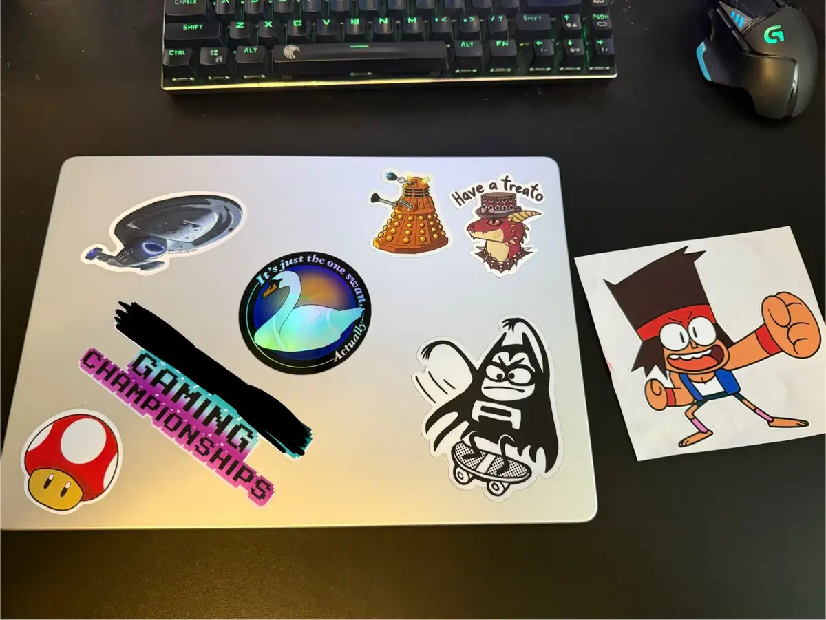 A silver laptop lid covered in various colorful stickers including a Dalek, a swan with text, a Mario mushroom, and a…
