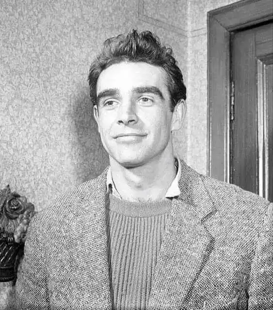A black-and-white photo of a man with styled hair, wearing a textured blazer over a ribbed sweater, standing indoors near a…