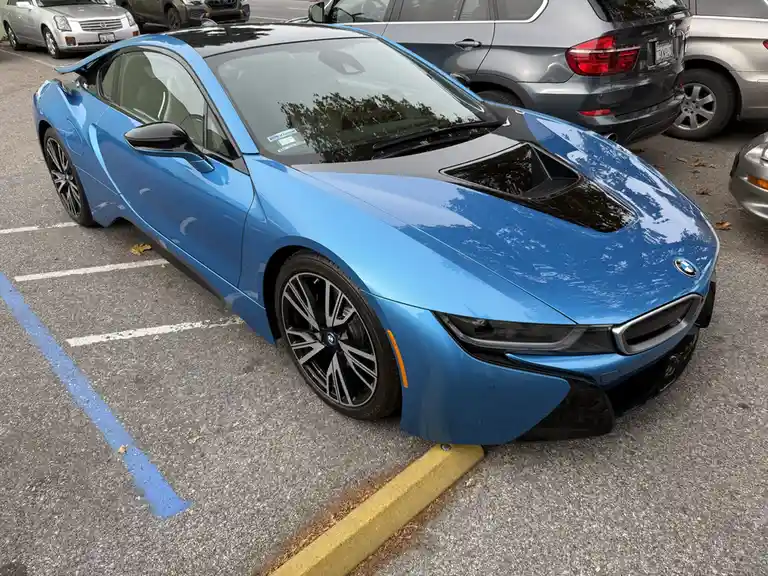 A blue BMW i8 sports car is parked in a parking lot, angled slightly toward the viewer, with its front bumper touching a…