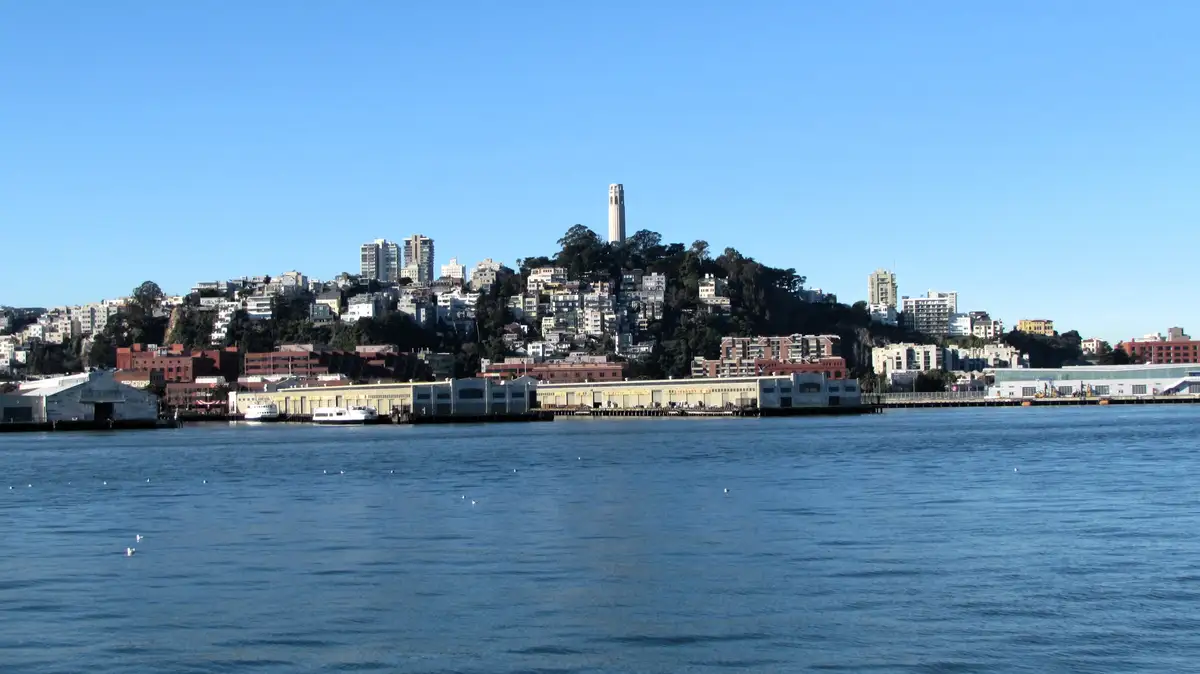 A scenic view of the San Francisco waterfront with Coit Tower visible atop a hill, surrounded by city buildings and water…