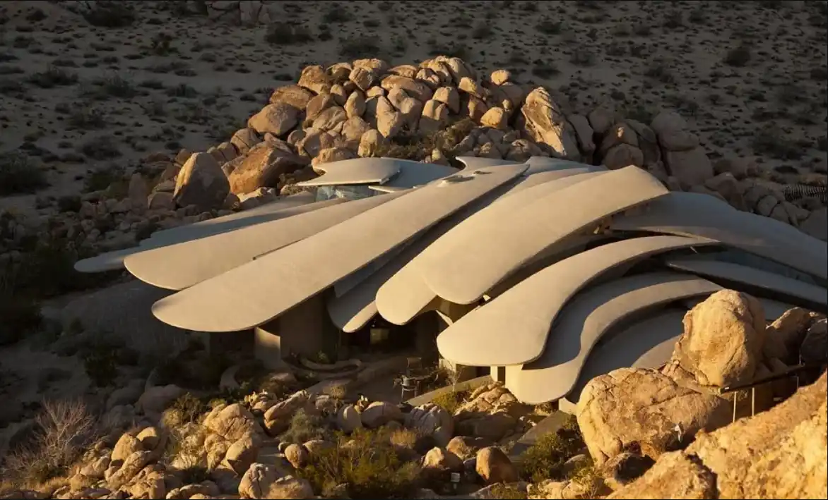 an aerial view of a modern, sculptural house with sweeping, wing-like concrete roofs nestled among large desert boulders and…
