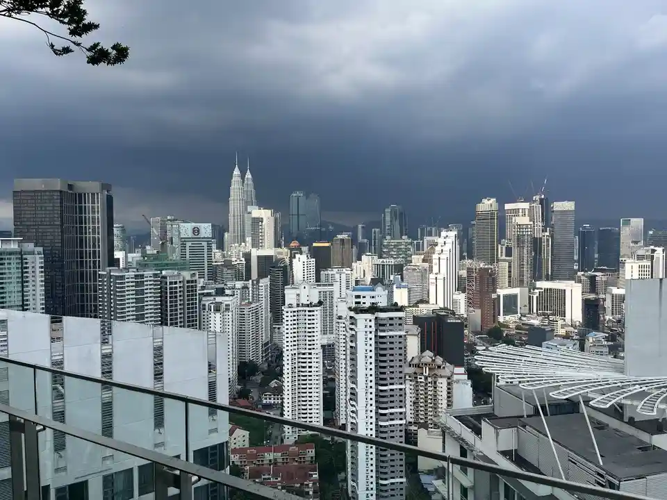 A cityscape photo of Kuala Lumpur with dark storm clouds looming over the skyline, featuring the Petronas Twin Towers and…
