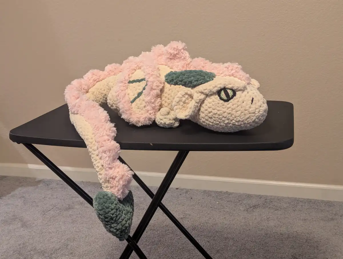 A handmade, fluffy crochet dragon plushie with pink and cream coloring and green eyes is lying on a black folding table…