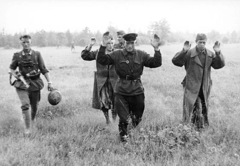 A black-and-white historical photograph shows several men in military-style clothing walking through a grassy field, all…