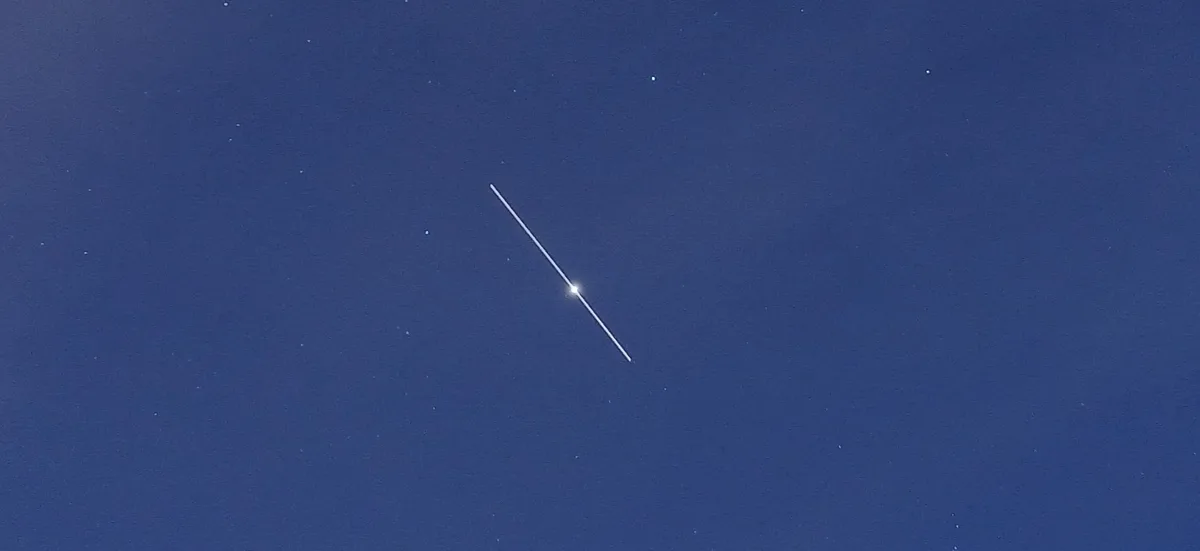 A dark blue night sky with faint stars, featuring a bright, diagonal white streak with a glowing point at its center…