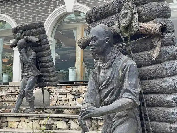 Two bronze statues of men carrying heavy loads of logs on their backs stand on stone steps. The man in the foreground looks…