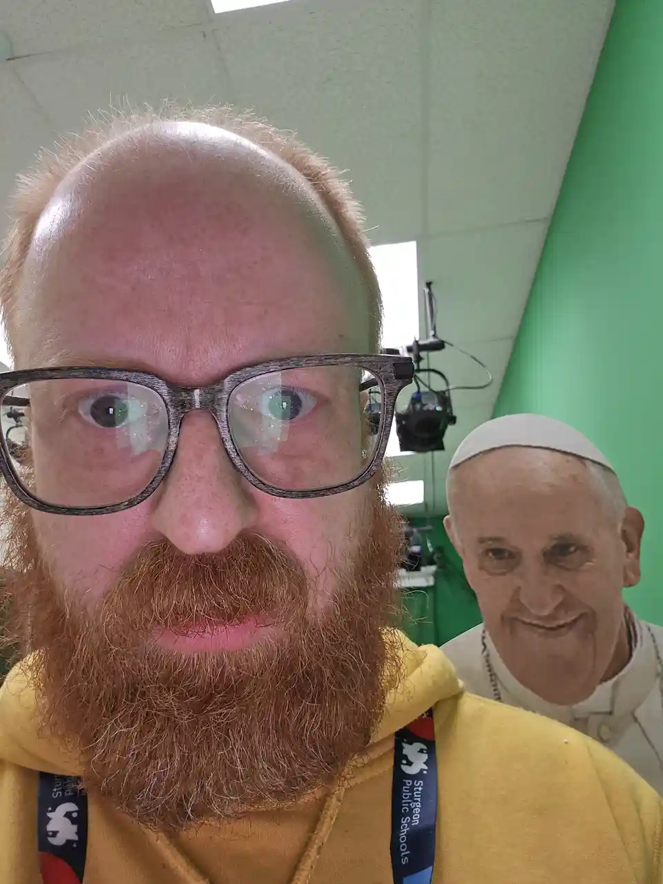 A selfie of a man with a red beard and glasses wearing a yellow hoodie, with a cutout image of Pope Francis superimposed on…