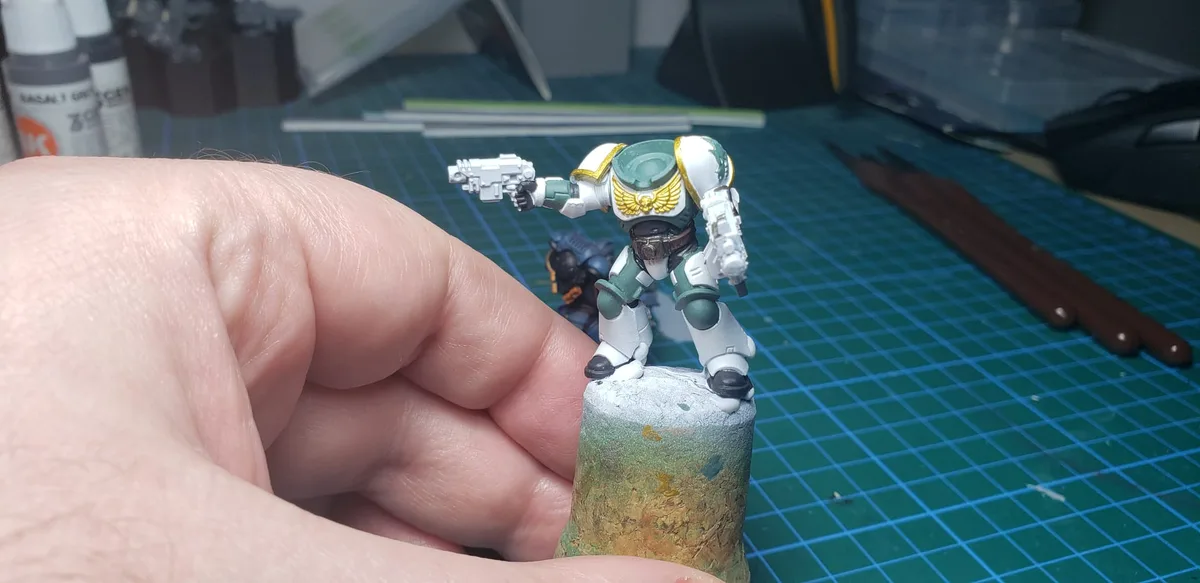 A hand holds a painted miniature sci-fi warrior figure in green and white armor with gold wings, standing on a textured…
