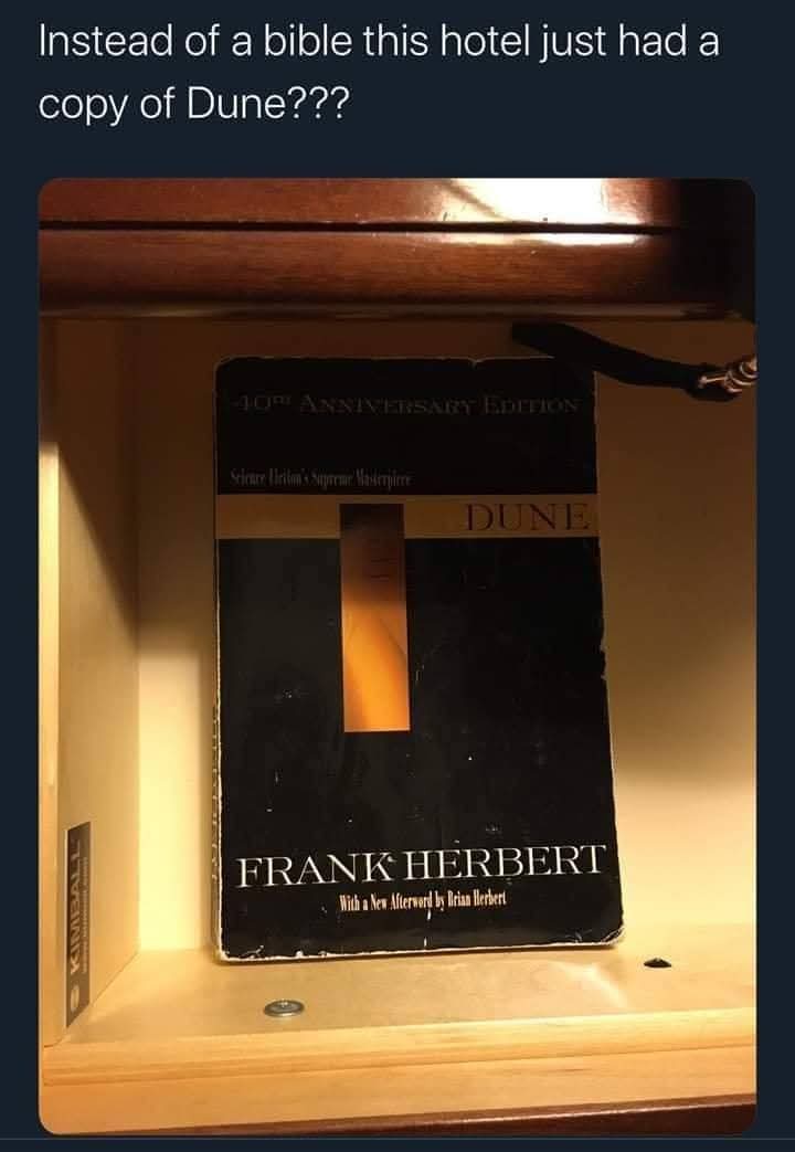 A close-up photo shows a worn copy of Frank Herbert's 'Dune' (40th Anniversary Edition) sitting on a wooden shelf, likely in…
