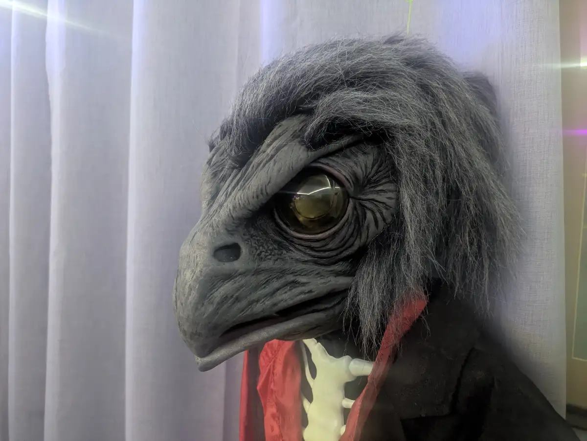 A close-up of a grey, bird-like creature mask with detailed feathers, a large golden eye, and a slightly open beak. The mask…