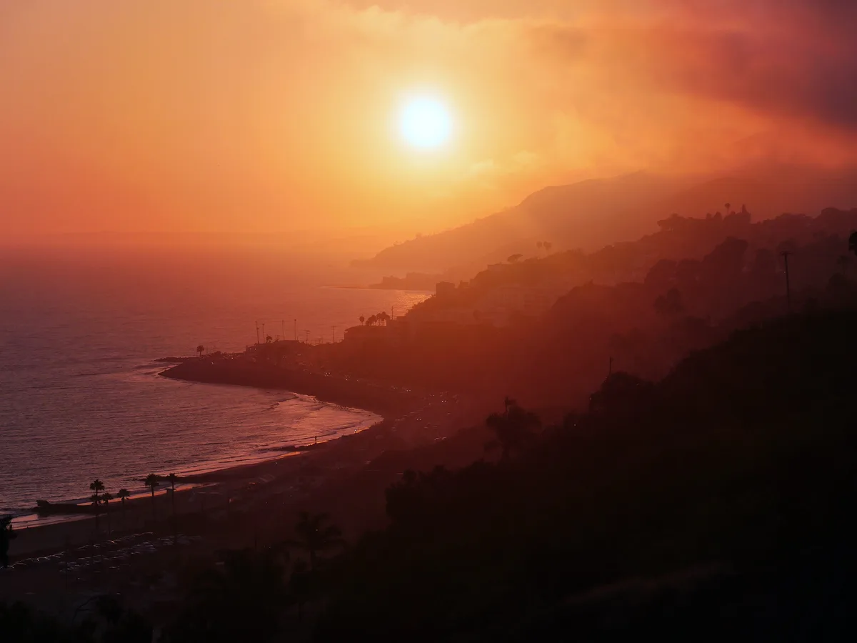 A scenic coastal sunset with a bright sun, orange sky, and silhouetted hills, beach, and buildings. Overlaid text reads 'i'm…