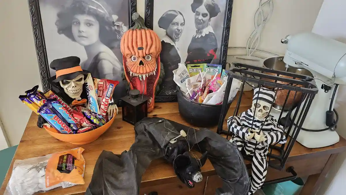 A wooden table is covered with Halloween decorations including a skeleton in a top hat holding a bowl of candy, a…