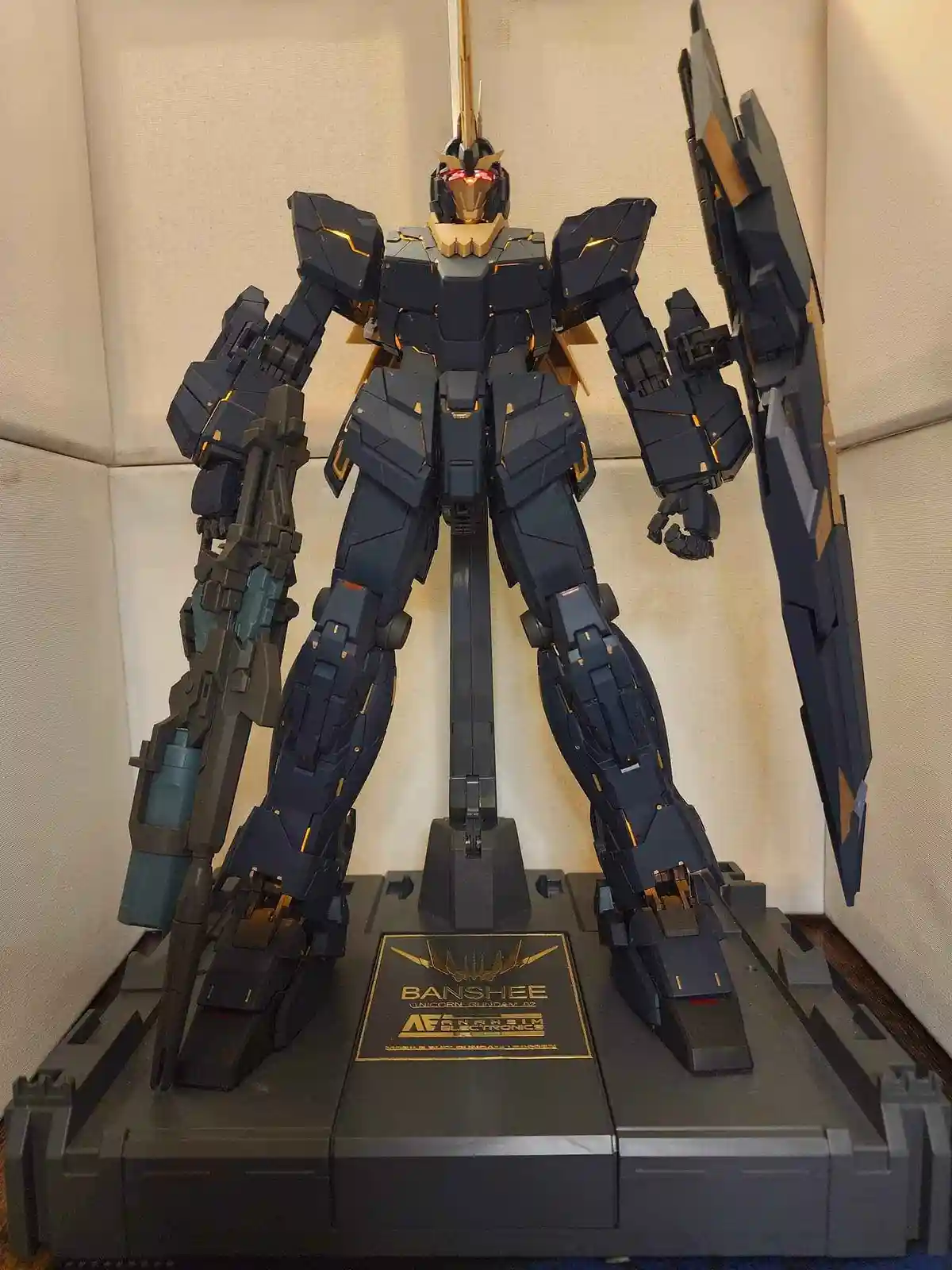 A detailed black and gold Gundam model, identified as the Banshee Unicorn Gundam A2, stands on a display stand holding a…