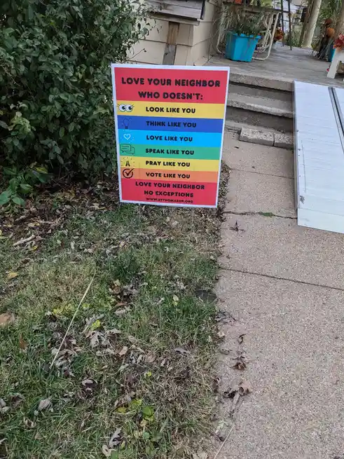 A colorful yard sign with a rainbow-striped background stands in grass next to a concrete sidewalk, listing conditions under…