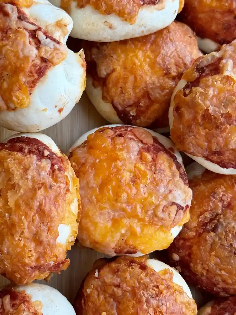 A close-up photo shows numerous small, round buns topped with melted orange cheese and red sauce, resembling miniature…