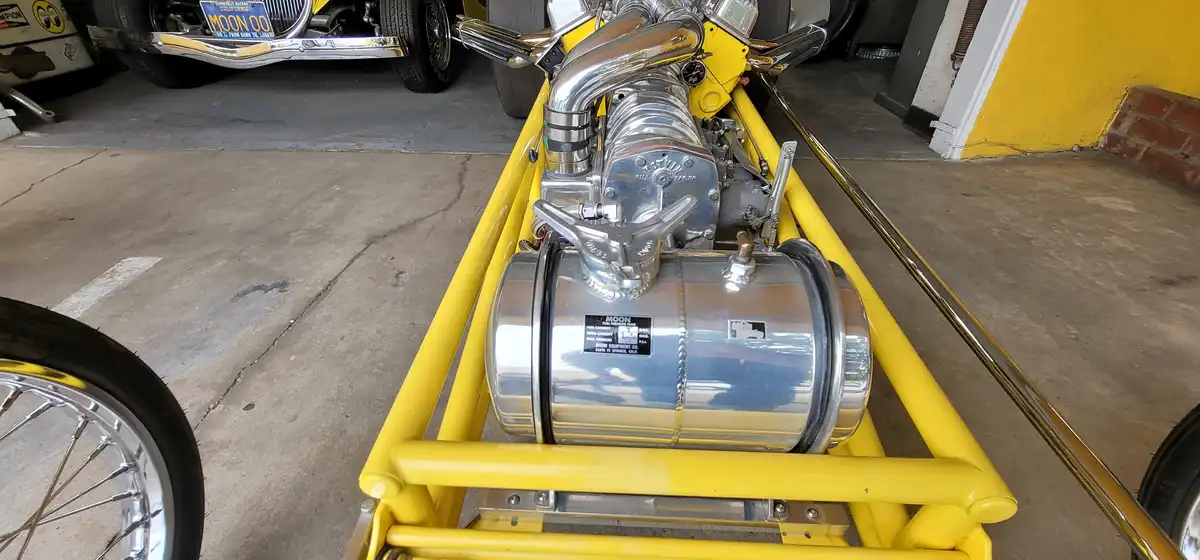 A close-up shot of a custom yellow hot rod's engine bay, featuring a shiny chrome fuel tank labeled 'MOON' and a matching…