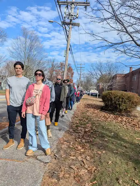 A long line of people stands on a sidewalk next to a grassy area with fallen leaves under a partly cloudy sky. In the…