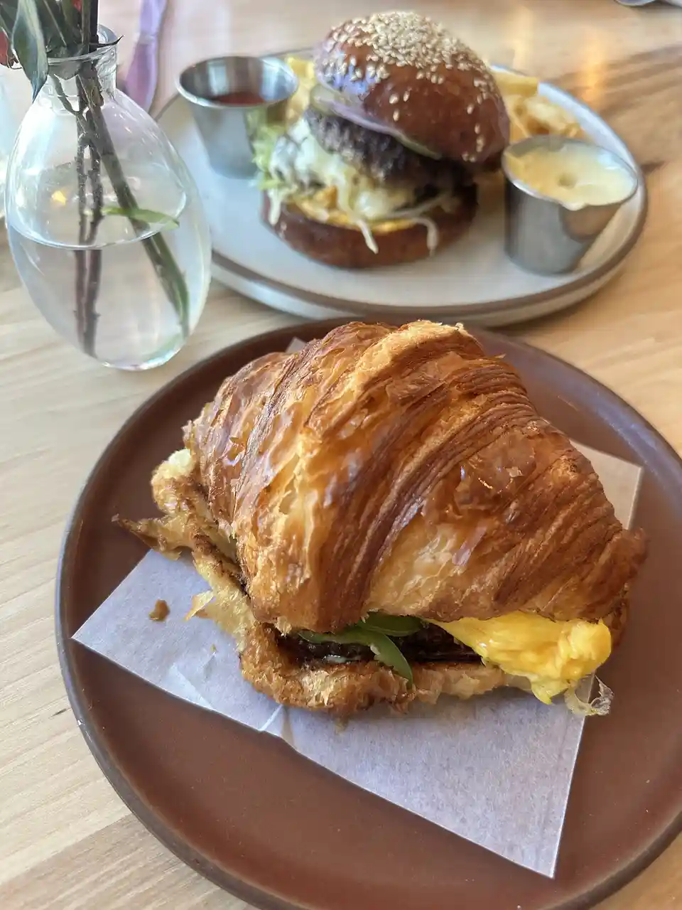A close-up photo shows a golden-brown croissant sandwich on a brown plate in the foreground, with a classic burger and fries…